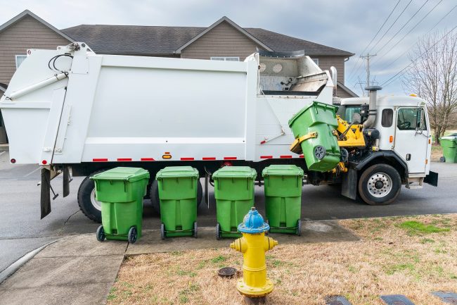 Horizontal shot of an automated garbage truck picking up trash container on curb.  Four more garbage cans are lined up waiting their turn.